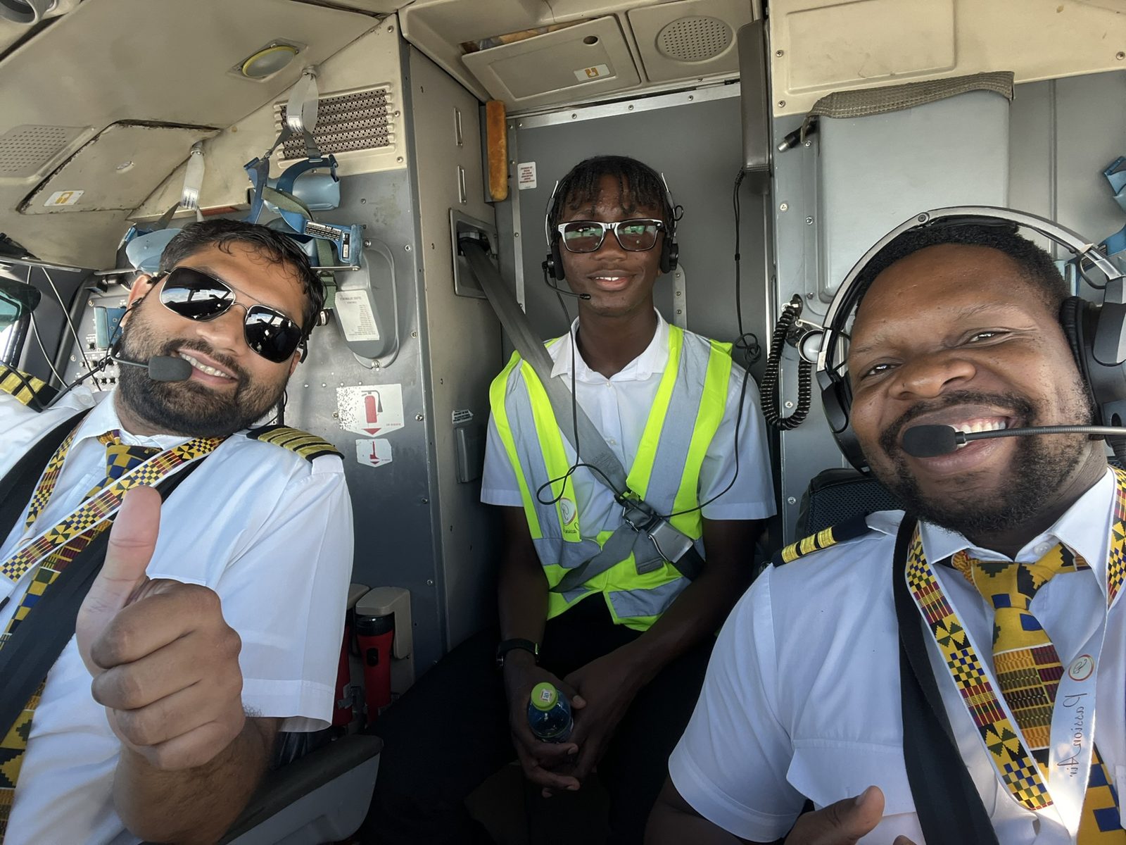 Kobi in a hi-vis vest on the flight deck jump seat between two smiling Passion Air pilots