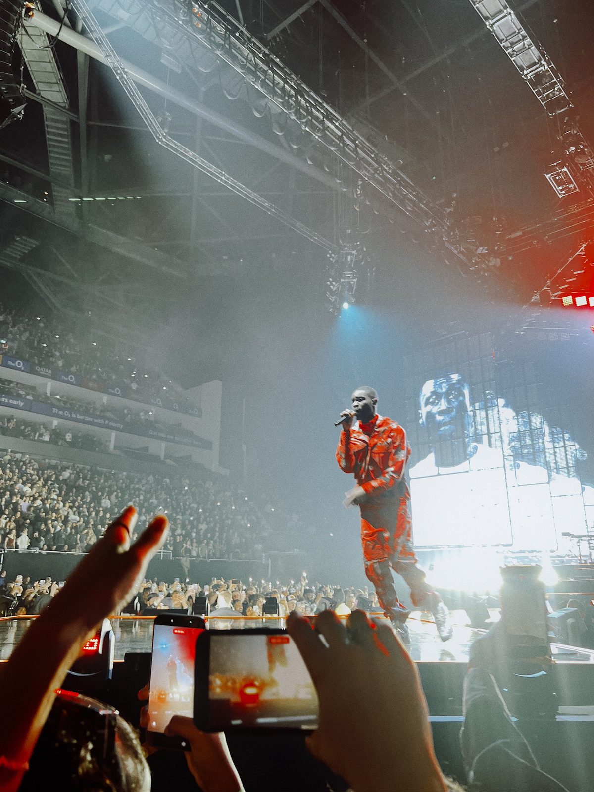 Dave in a red suit on stage at the O2, phones raised in the crowd